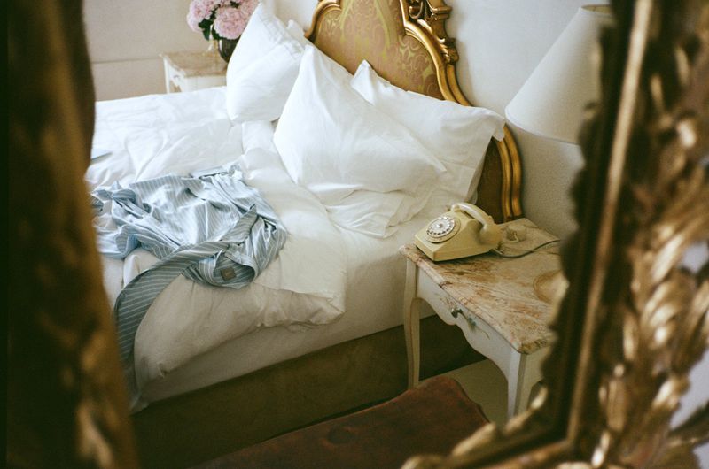 Bedroom scene with an unmade bed, white linens, gold headboard, vintage phone, and hints of light in a domestic interior.