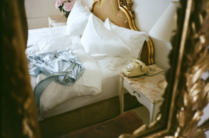 Bedroom scene with an unmade bed, white linens, gold headboard, vintage phone, and hints of light in a domestic interior.