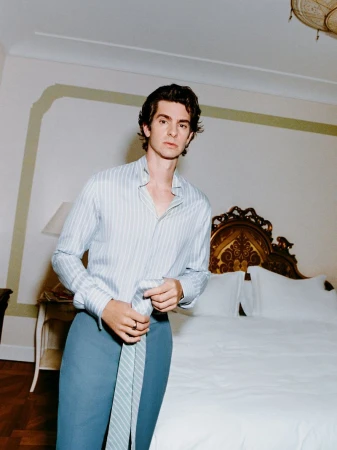 Portrait of Andrew Garfield indoors, wearing a striped shirt and blue pants, loosely holding a striped tie, in a hotel-like setting.