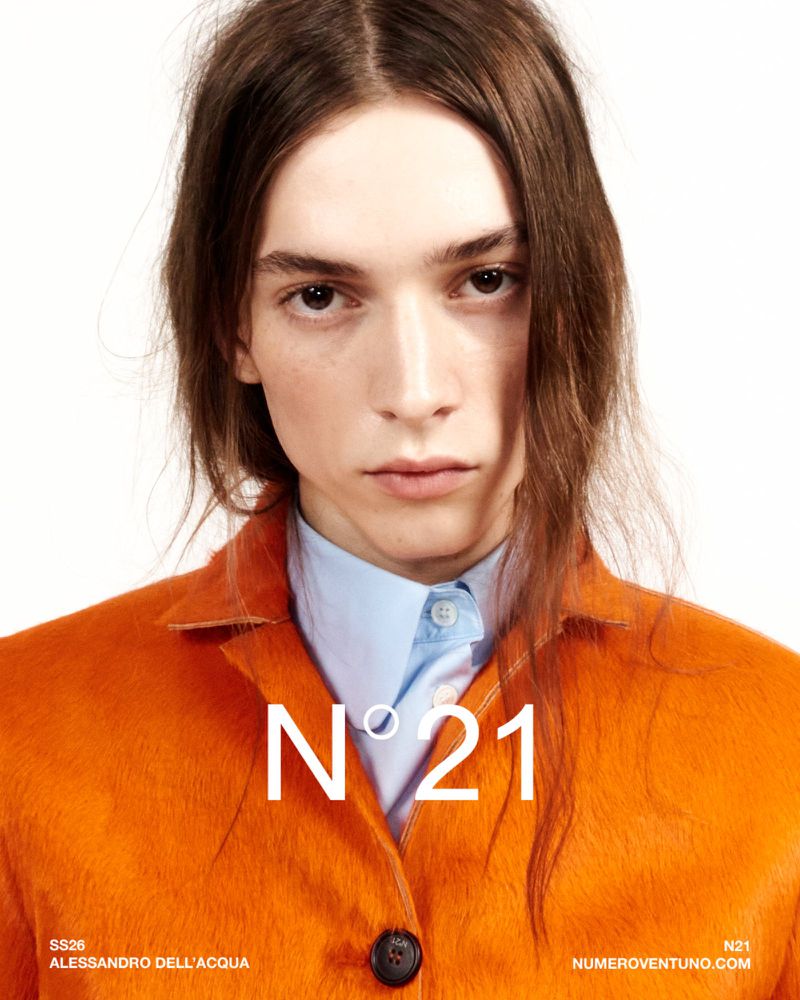 Close-up portrait of a model with long brown hair wearing a bright orange textured jacket and blue shirt against a white backdrop.