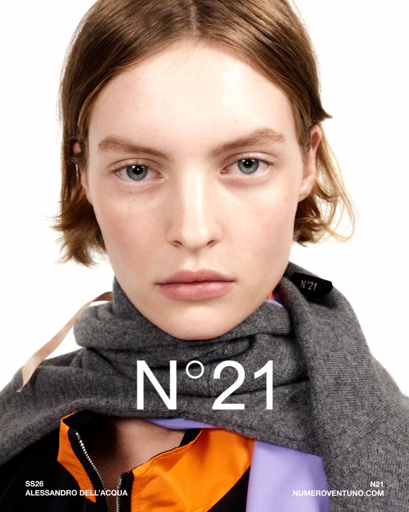 Close-up portrait of a fair-skinned adult female wearing a gray scarf and a designer jacket against a stark white backdrop, fashion campaign.
