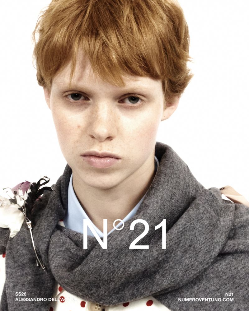 Close-up fashion shot featuring a young adult with red hair wearing a grey scarf and floral accents on a white background.