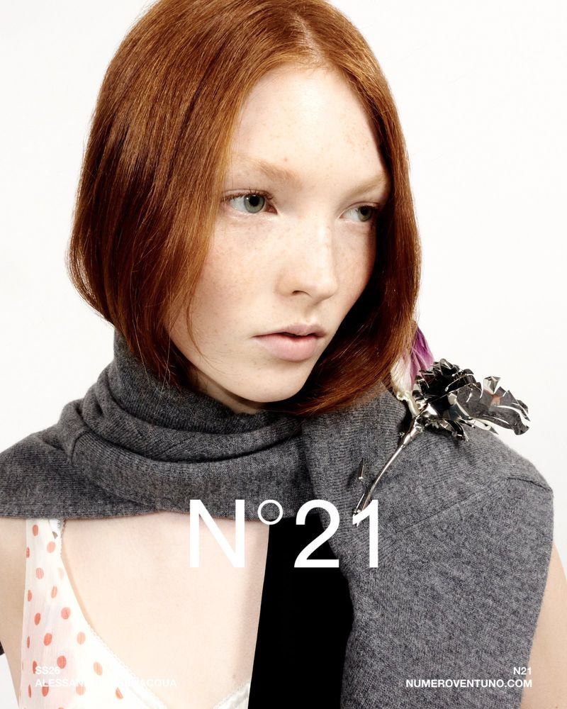 Close-up studio shot of a red-haired woman wearing a grey scarf and rose brooch against a white background with fashion logo text overlay.