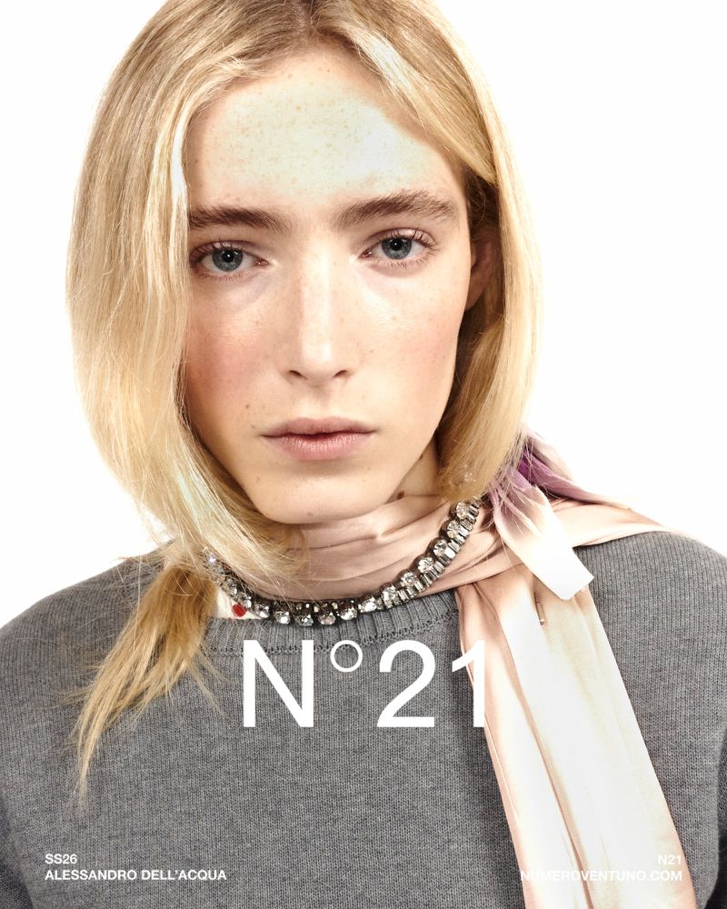 Close-up fashion shot of a blonde woman wearing a grey sweater and rhinestone necklace against a white background with N21 text overlay.