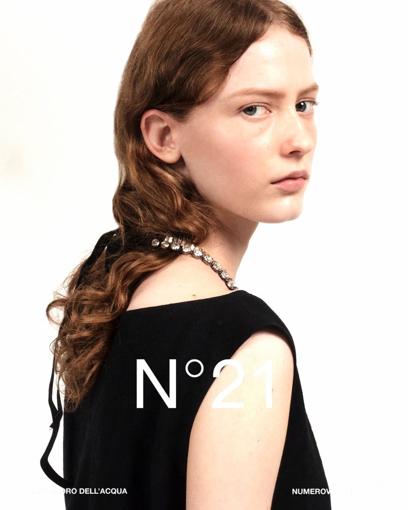 Close-up studio shot of a young woman with red hair wearing a black dress and rhinestone necklace against a white background. 