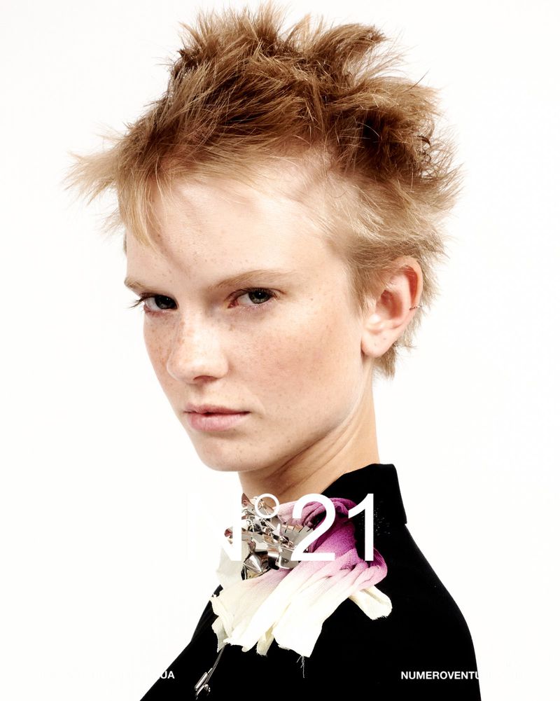 Close-up shot of a fair-skinned young woman with short, spiky blonde hair wearing a black top with a floral collar embellishment.