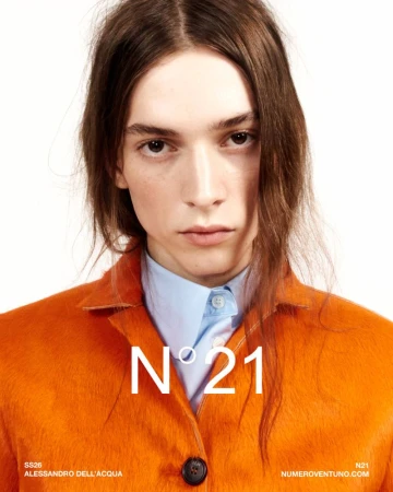 Close-up portrait of a model with long brown hair wearing a bright orange textured jacket and blue shirt against a white backdrop.
