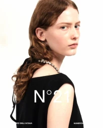 A model with wavy brown hair looks over her shoulder wearing a black sleeveless top with a rhinestone strap against a white backdrop.