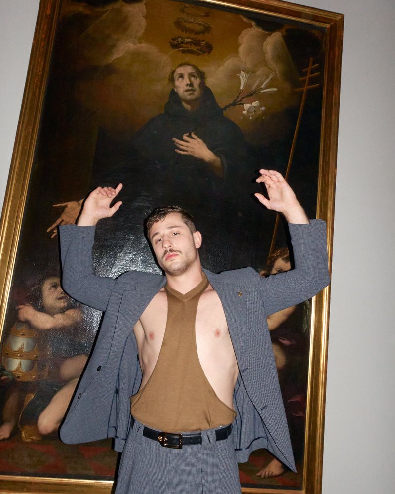 Adult male posing in front of framed religious artwork with expressive gesture, wearing grey suit, open brown top and black belt indoors.