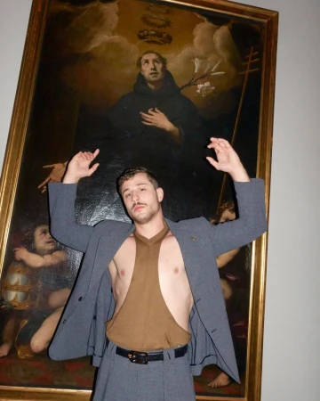 Adult male posing in front of framed religious artwork with expressive gesture, wearing grey suit, open brown top and black belt indoors.