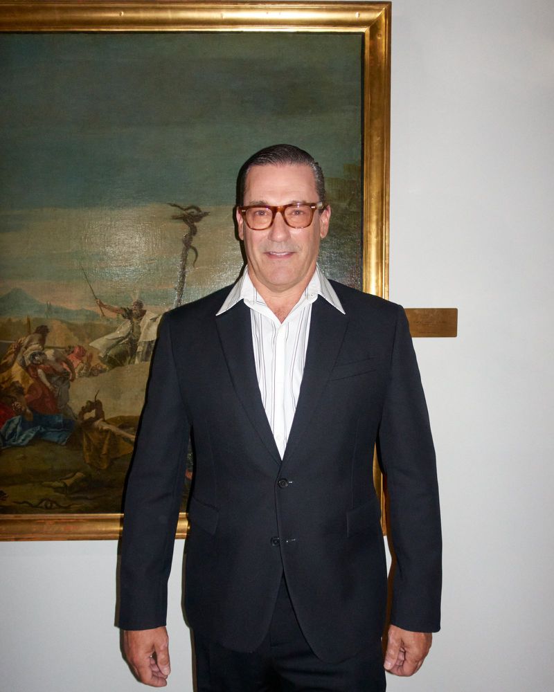 Adult male in dark suit and glasses stands against a white wall with a large landscape painting in a gold frame.