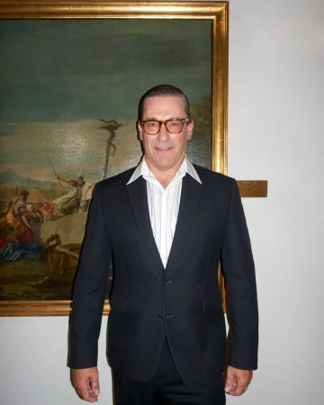 Adult male in dark suit and glasses stands against a white wall with a large landscape painting in a gold frame.
