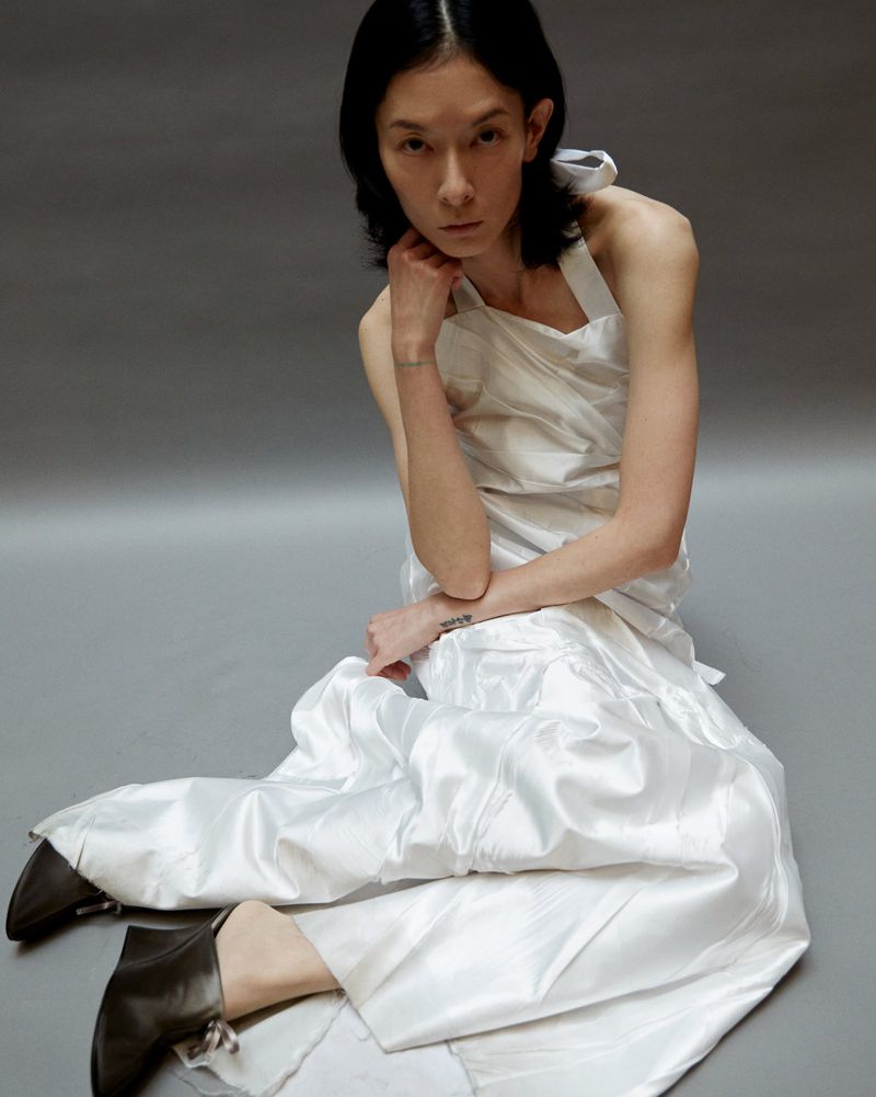 A fashionable person with dark hair poses in a white halter top and wide-legged satin pants, seated against a grey studio backdrop, looking at camera.