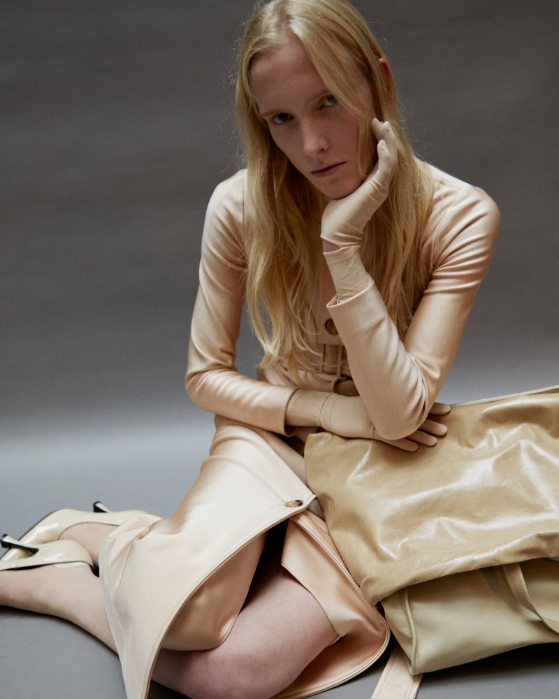 A blonde model poses indoors in a beige dress, matching gloves, and high heels, next to a beige leather bag on a gray background.
