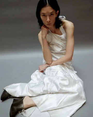 Model Hsin-Yu Liu seated on a grey studio floor wearing a white draped satin halter dress and black pointed mules.
