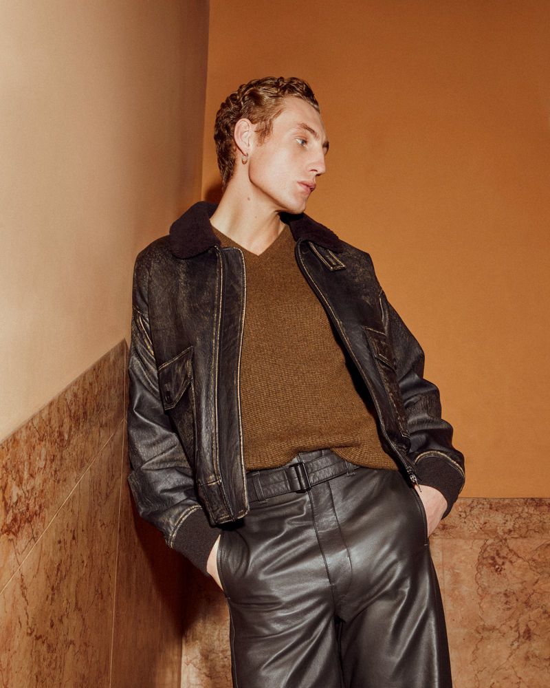 Adult male fashion model in a brown sweater, leather jacket, and black pants posing indoors with hands in pockets looking off-camera.