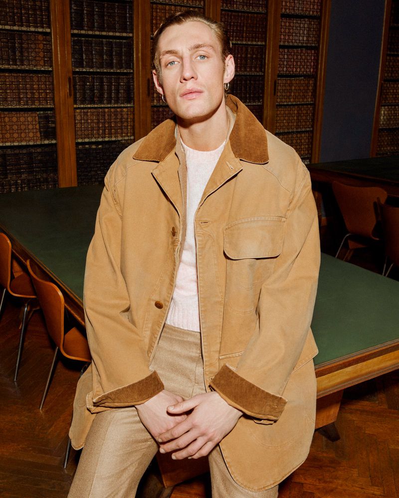 Aubrey O'Mahony wearing a tan corduroy-collared barn jacket and beige trousers seated by a library table.