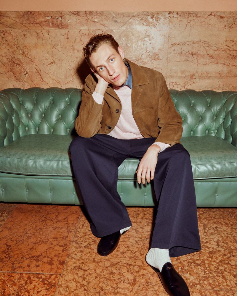 Young man lounges on a green tufted couch, wearing a brown suede jacket and navy trousers, set against a textured peach wall.
