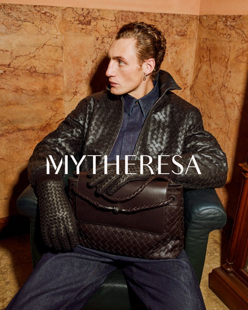Stylish man in a black woven jacket and dark jeans sits in an armchair holding a brown woven handbag in a warm indoor setting.