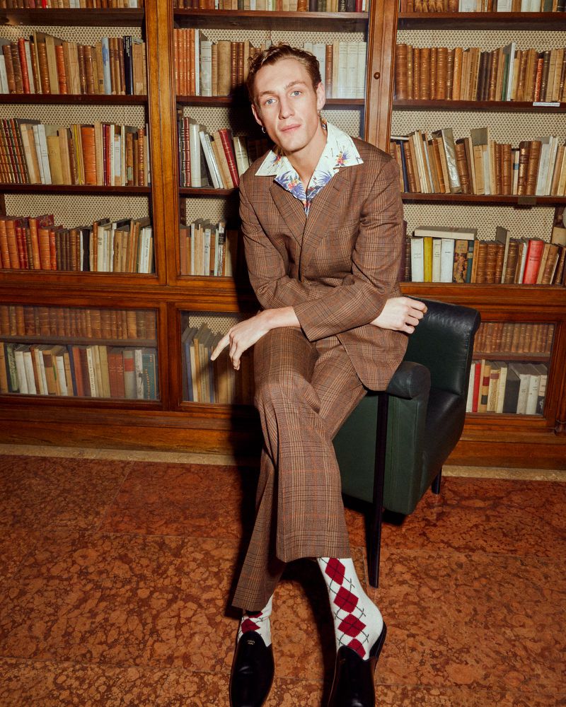 A fair-skinned adult male in a brown suit poses while seated in a green chair in front of a bookcase filled with books inside.
