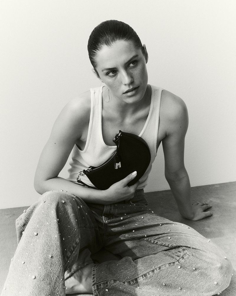 Model Emma Todt seated on the floor holding a black crescent bag, wearing a white tank top and rhinestone-embellished jeans.