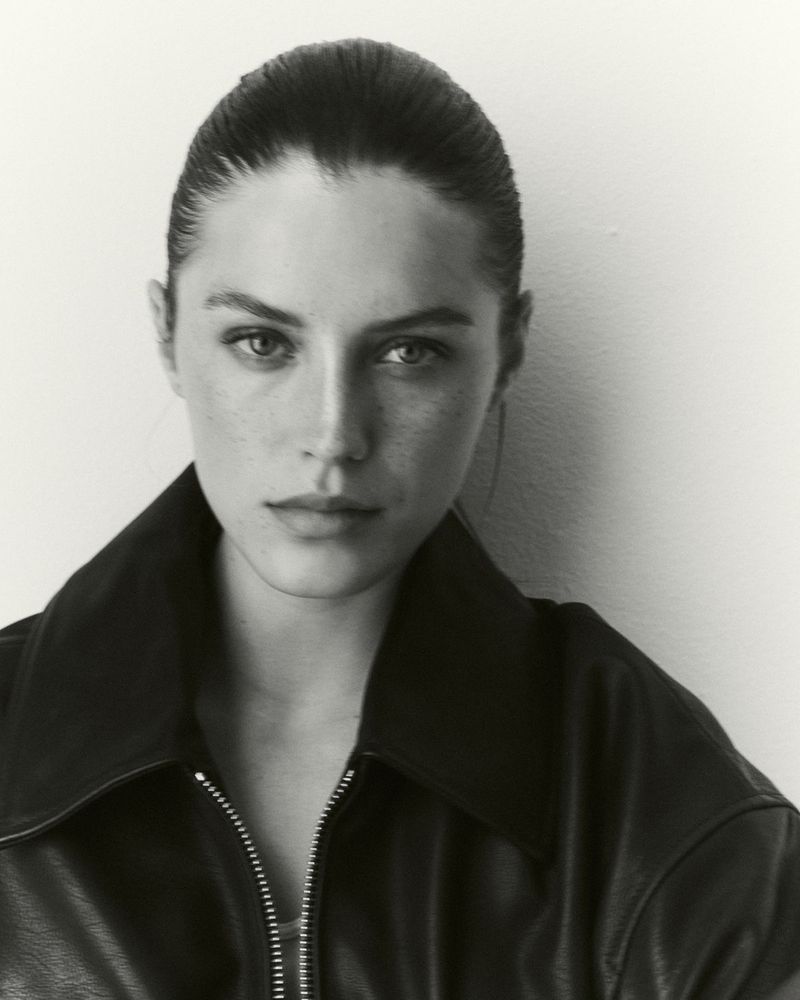 Close-up portrait of a young woman with slicked-back hair, wearing a leather jacket in a brightly lit, neutral setting.