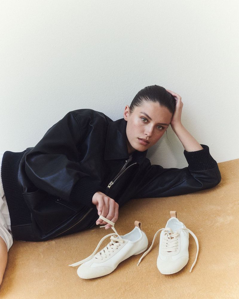 Woman poses with white sneakers indoors against a two-tone background of white and tan, wearing a black jacket and casual clothing.