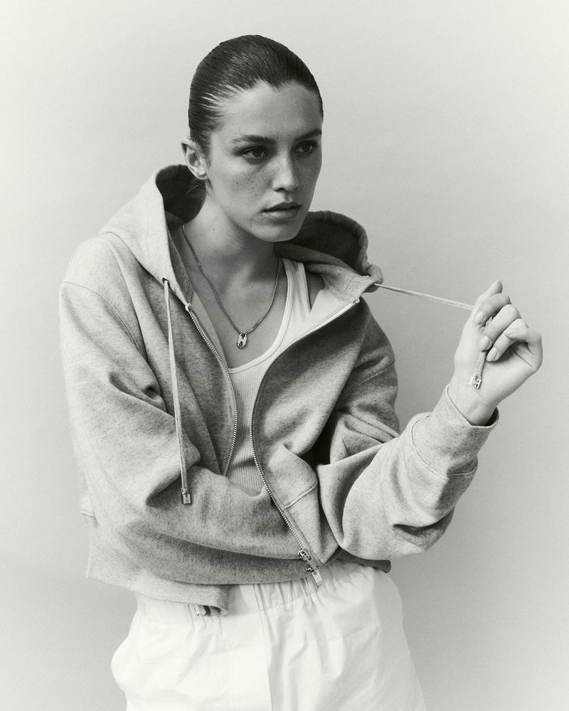 Monochrome portrait of a young adult female wearing a gray zip-up hoodie and white pants, showcasing a necklace and holding a drawstring.