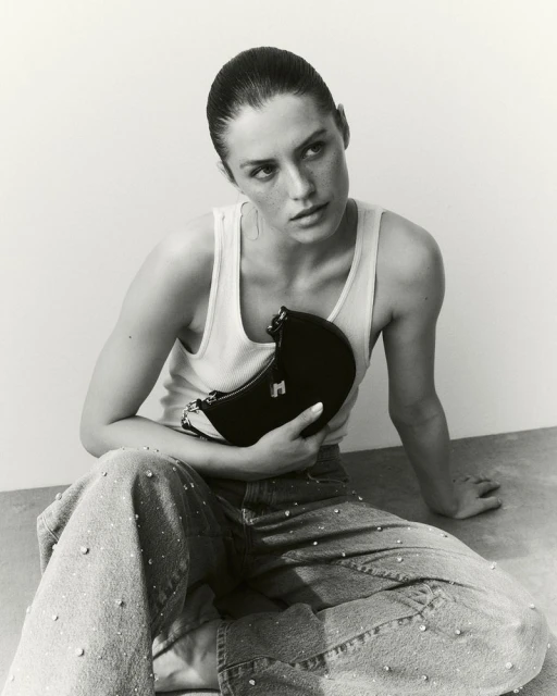 Model Emma Todt seated on the floor holding a black crescent bag, wearing a white tank top and rhinestone-embellished jeans.