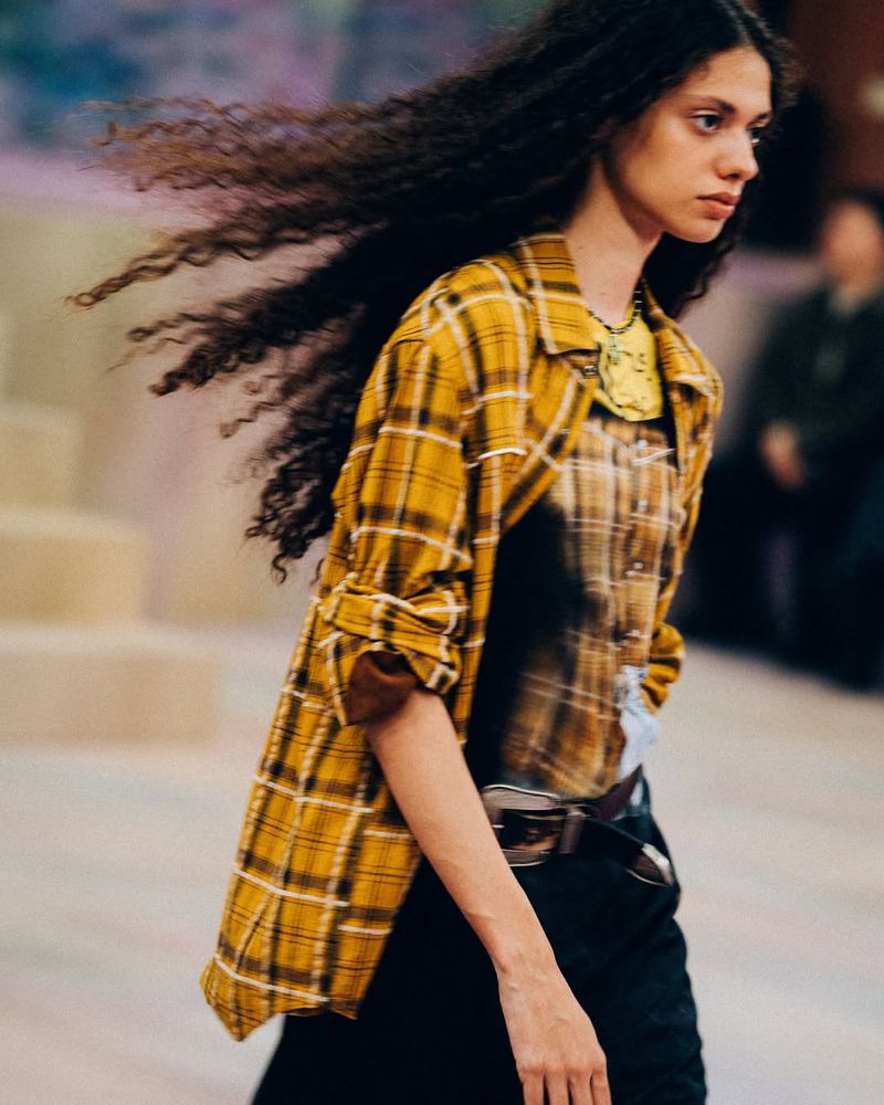 A young woman with dark curly hair is walking. She is wearing a yellow plaid shirt, dark pants, and a brown belt in an indoor setting.