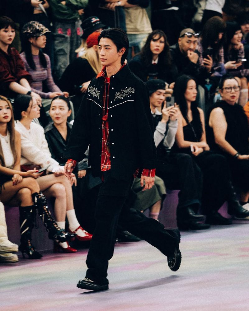 Fashion show featuring a runway and a male model in a black jacket, red shirt, and black pants walking in front of a seated audience.