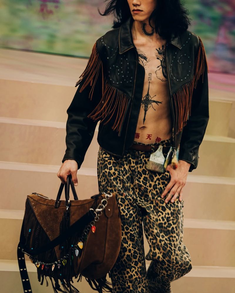 A person walks down stairs wearing a leather fringe jacket, leopard print pants, and carrying a suede bag with fringe details and embellishments.