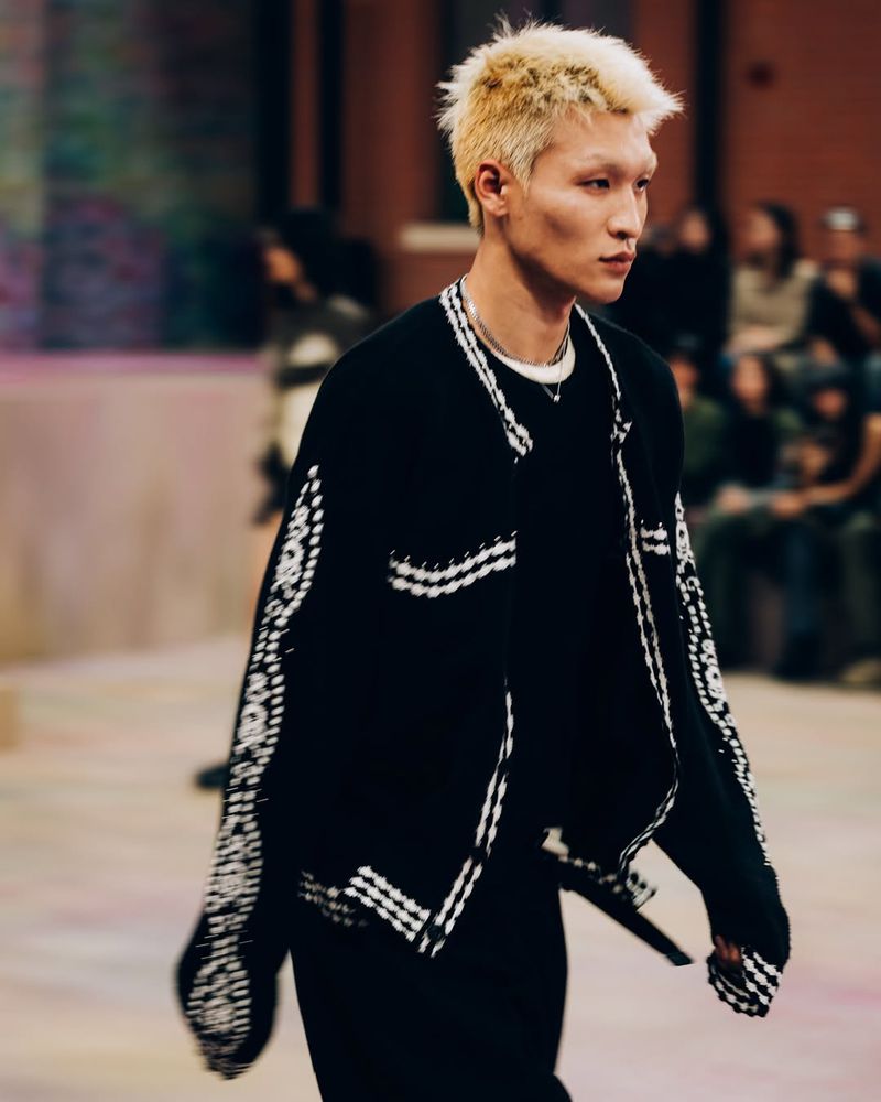 Runway shot of a fashion model in black patterned jacket with short, blond hair, wearing a black shirt and pants, with audience seating in background.
