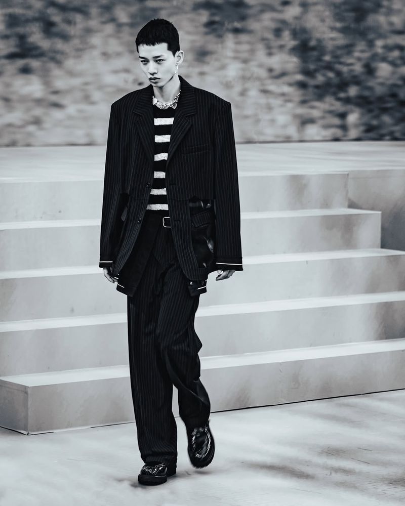 Monochrome portrait of a male fashion model on a runway, showcasing an oversized pinstripe suit and striped shirt during a show.