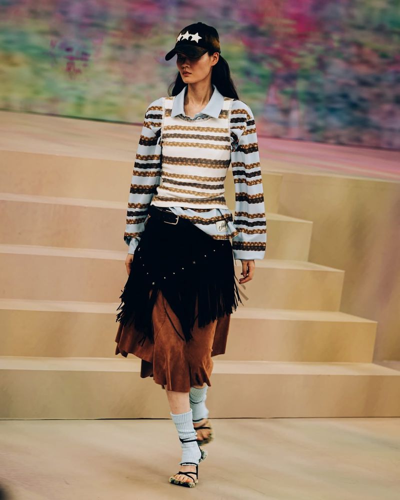 Runway model walks the catwalk wearing a striped top, layered skirt, socks, and sandal heels, accessorized with a star-studded baseball cap.