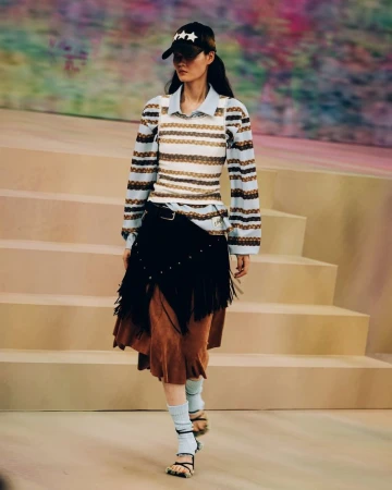 Runway model walks the catwalk wearing a striped top, layered skirt, socks, and sandal heels, accessorized with a star-studded baseball cap.