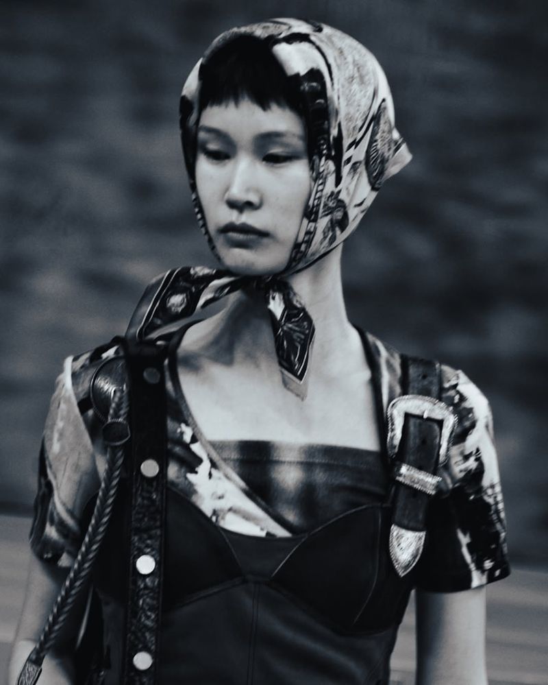 Monochrome fashion shot of an Asian woman wearing a patterned headscarf and top, with leather accents and a blurred background.