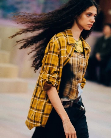 Model with long dark hair walking the runway wearing a yellow plaid shirt over layered tops for ADSB Andersson Bell