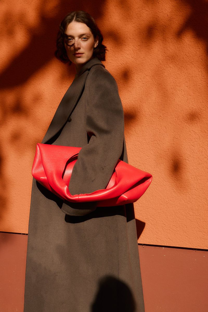 An adult woman stands against an orange wall, holding a large, vibrant red handbag. She is wearing a dark grey coat.