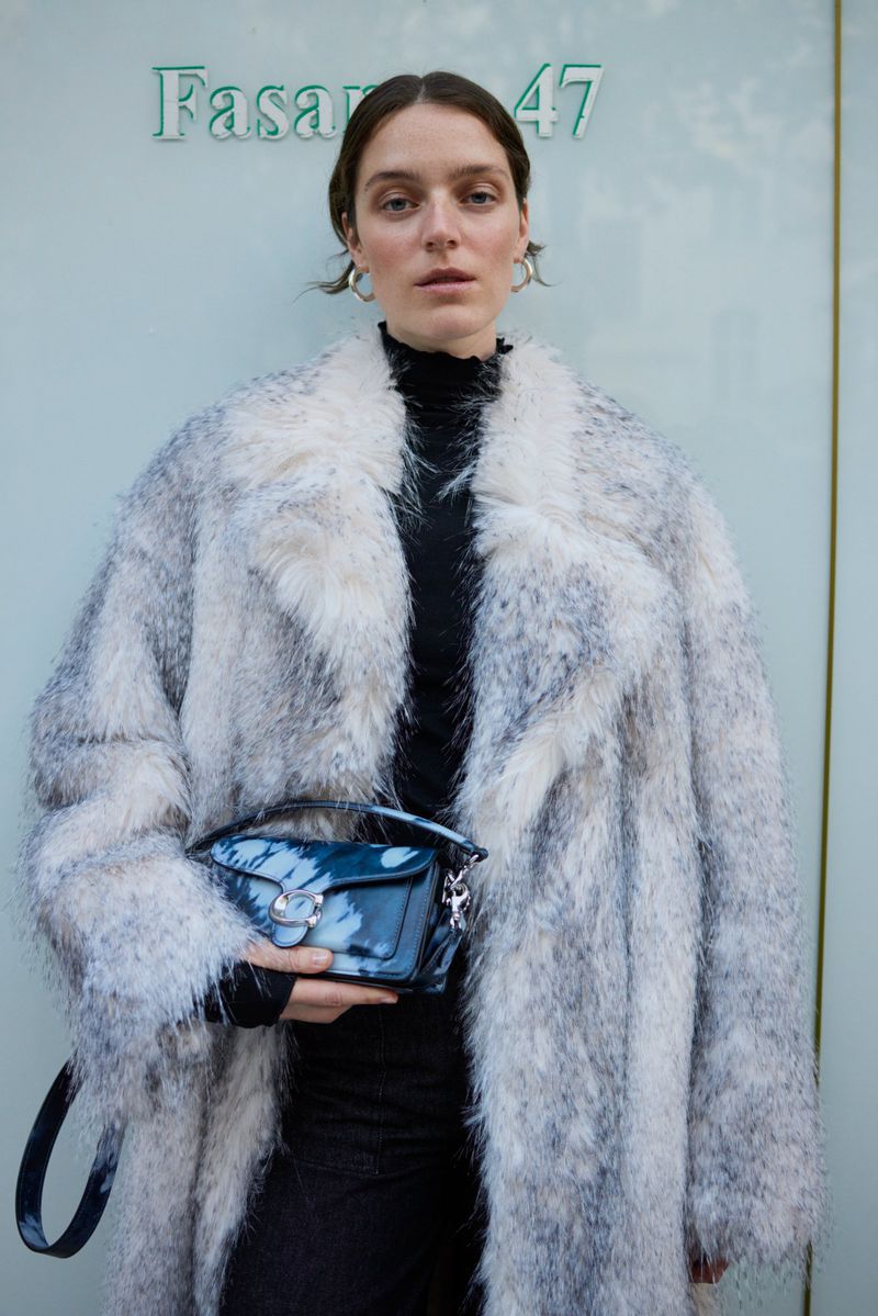 Adult woman in a fuzzy grey-white coat holds a blue tie-dye purse in front of a light blue building with green text signage.