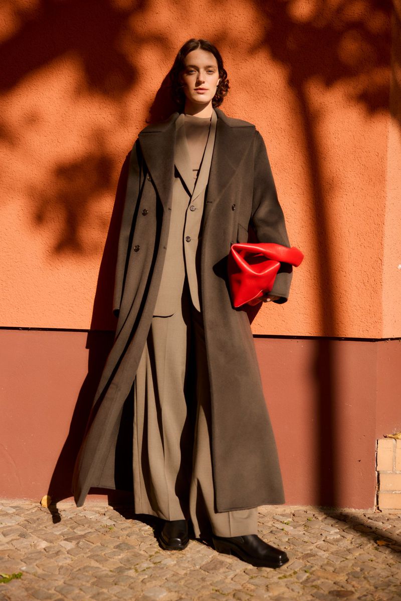 Fashionable adult female in a long dark coat and wide-legged pants poses against an orange wall with dappled tree shadows.