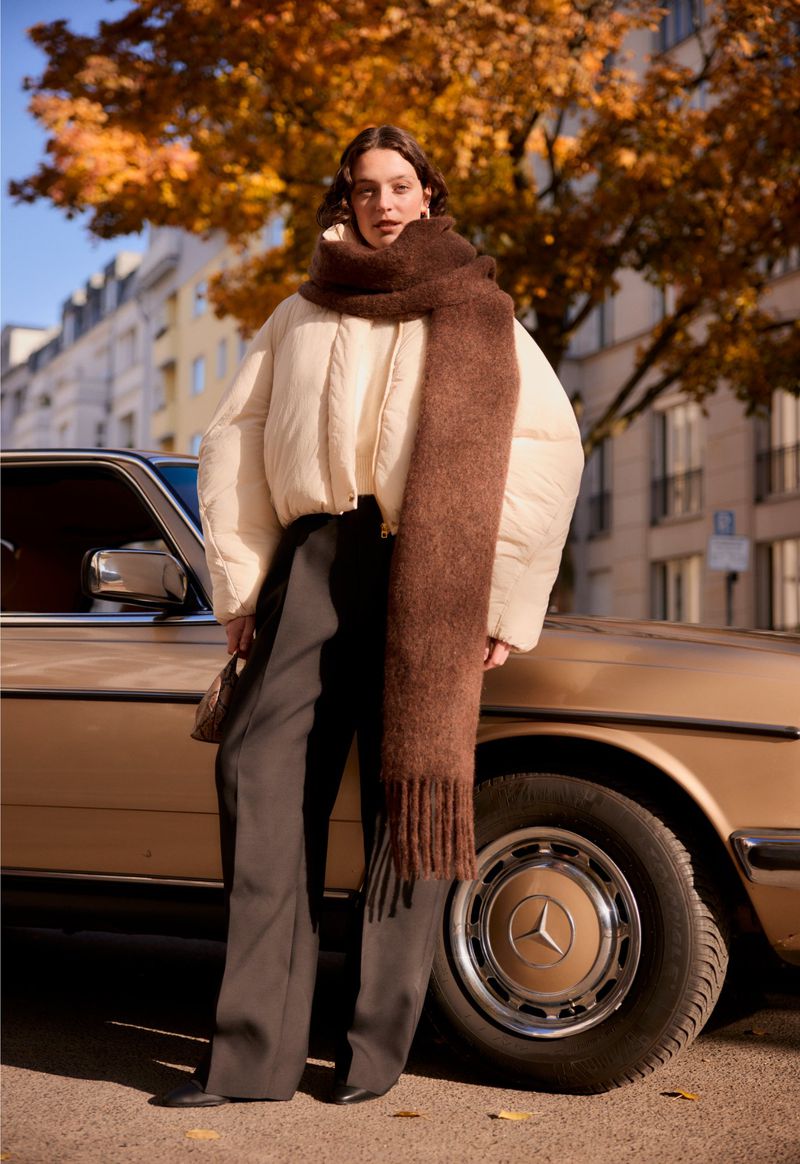Stylish adult female in a puffer jacket and scarf poses next to a vintage car on a city street with autumn trees in the background.