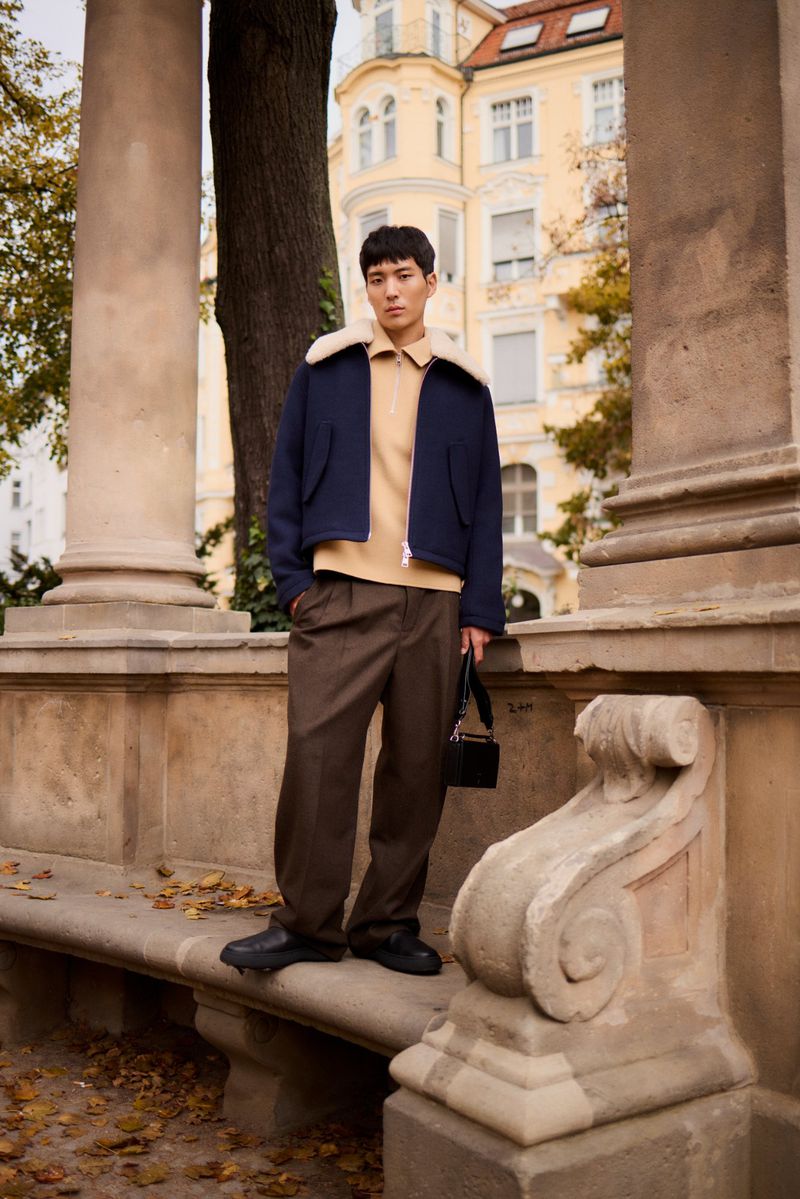Man in navy wool jacket with tan shirt and brown pants standing against stone columns in outdoor urban environment.