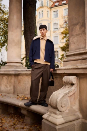 East Asian male model posing on a stone balustrade wearing a navy cropped jacket with shearling collar, tan knit, and brown trousers.