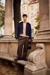 East Asian male model posing on a stone balustrade wearing a navy cropped jacket with shearling collar, tan knit, and brown trousers.
