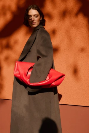 Woman in a long dark grey coat holding an oversized red leather clutch against a warm orange wall with dappled shadows.