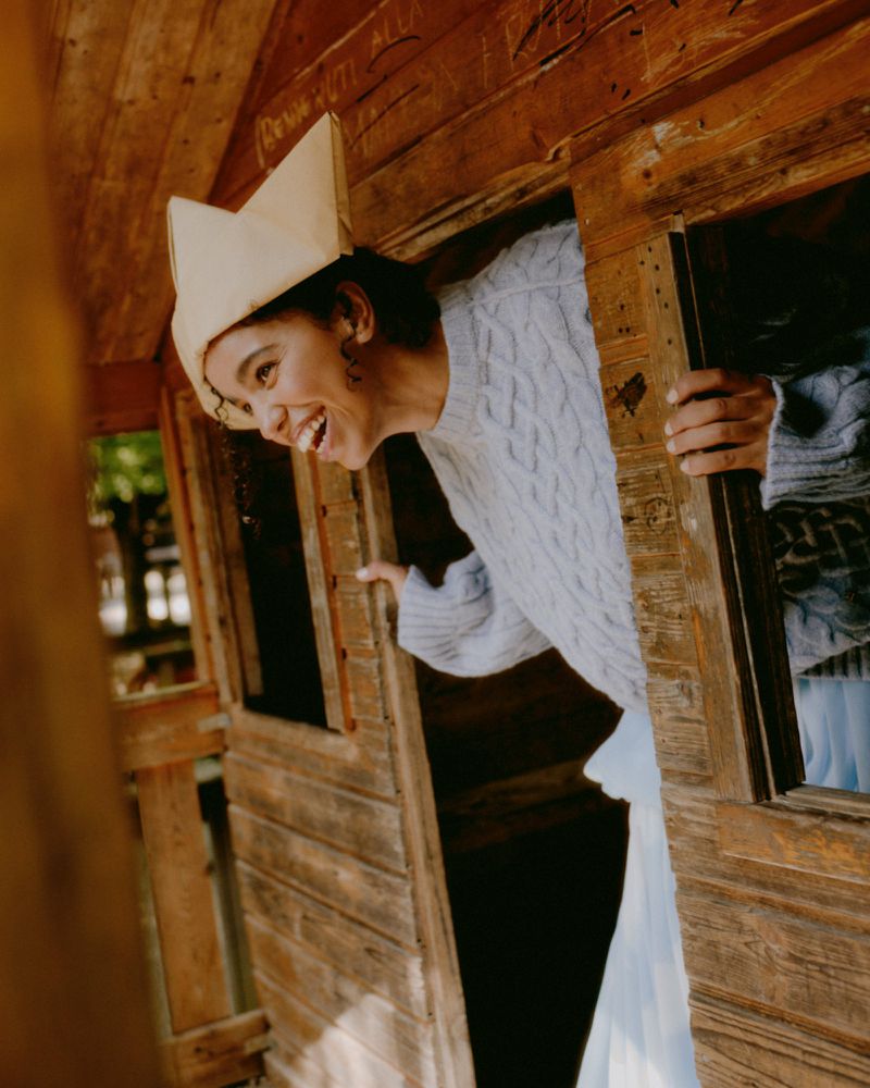 Young woman wearing a paper hat smiles, holding open a weathered wooden door of an outdoor playhouse on a sunny day in a park.