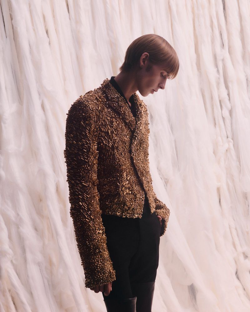 Adult fashion model poses in a textured metallic gold jacket against a draped white background in a studio setting with low lighting.