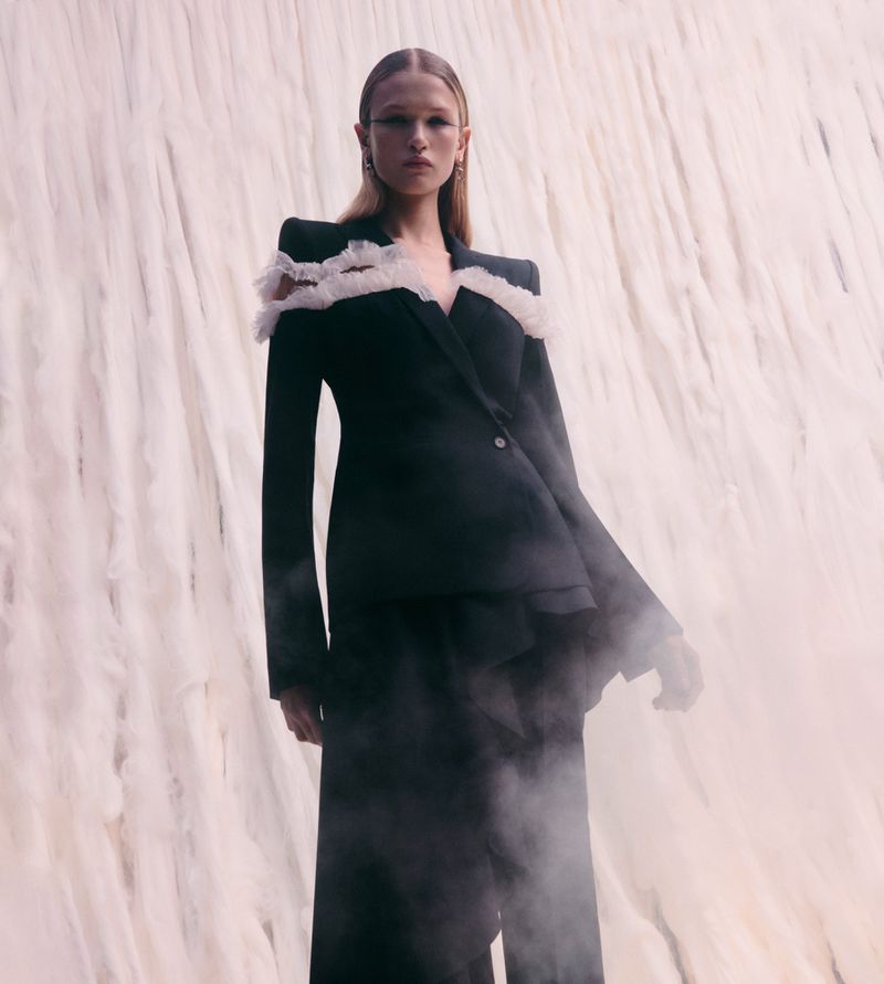 Woman in a black suit with white ruffle detail poses against a textured backdrop with smokey haze in a fashion shot.