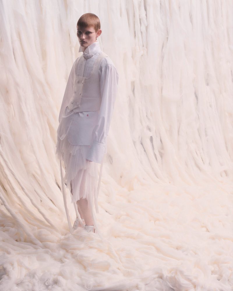 Fashion shot of a person in a white outfit on a soft, fluffy white surface against a draped backdrop, fashion-forward editorial style.